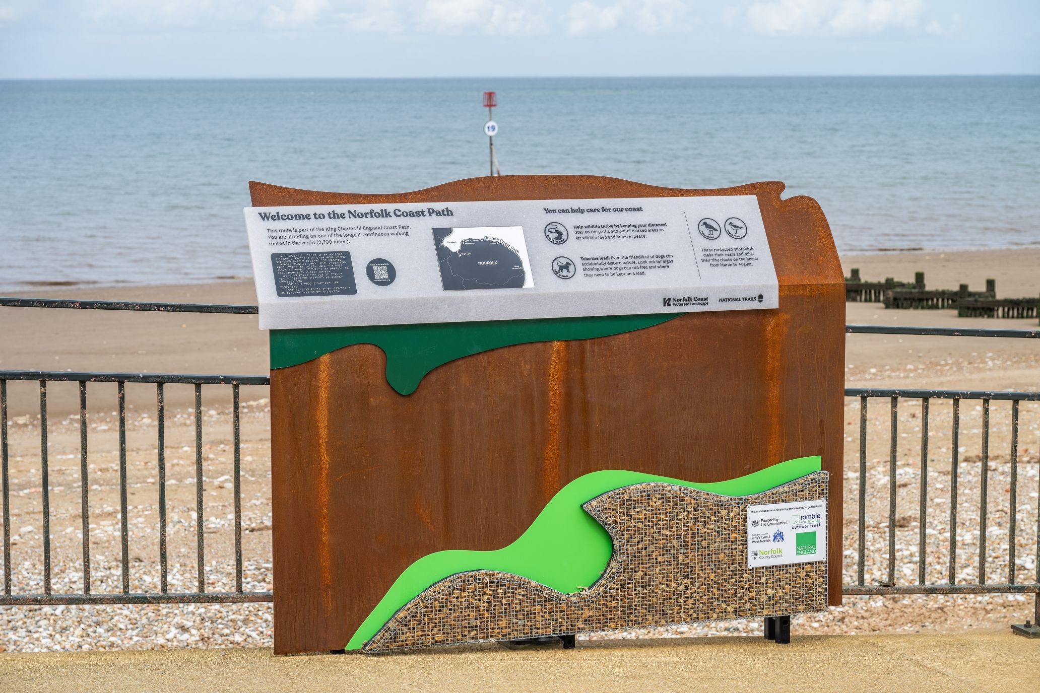 An interpretation panel along the walkway of a beach. This one is placed by the hand rail, looking out to sea. It includes text, braille and is placed on brassy metal. A small amount of shingle is placed on the bottom in front of a small, bright green, irregular shaped panel.