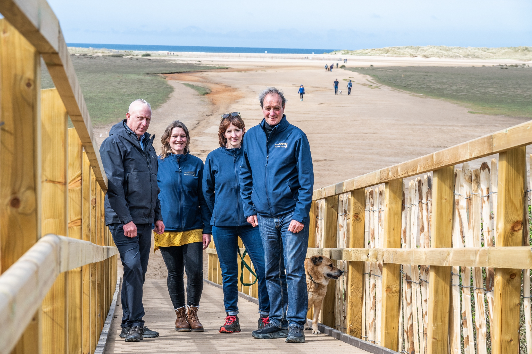 Jake Finnes, Manager of Farming at Holkham standing with Helen Terry, National Trail Team Leader, Katy Owen, Protected Landscapes Manager at Norfolk County Council and Cllr Andrew Jamieson, Chair of the Norfolk National Trail. These people all played in role in getting this boardwalk made. They are standing beside wooden handrails on the boardwalk, with Holkham beach in the background.
