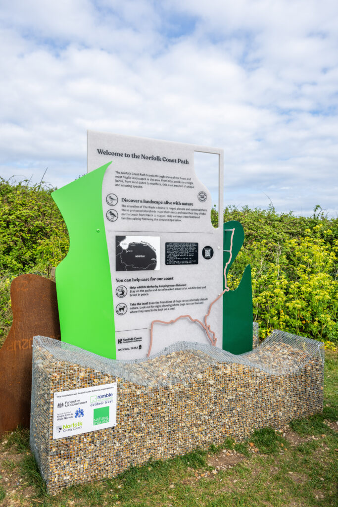 A new information board that has caged rocks on the bottom then irregular shapes in different colours, including brassy metal, bright green and dark green. The information board is grey and features a braille panel.