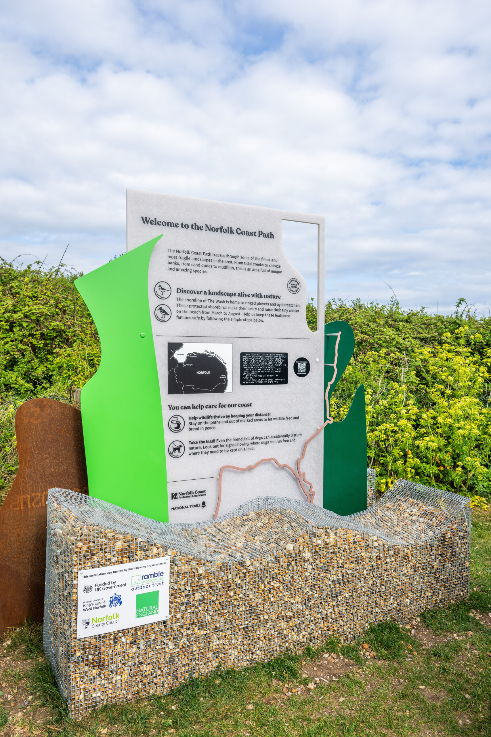 A new information board that has caged rocks on the bottom then irregular shapes in different colours, including brassy metal, bright green and dark green. The information board is grey and features a braille panel.
