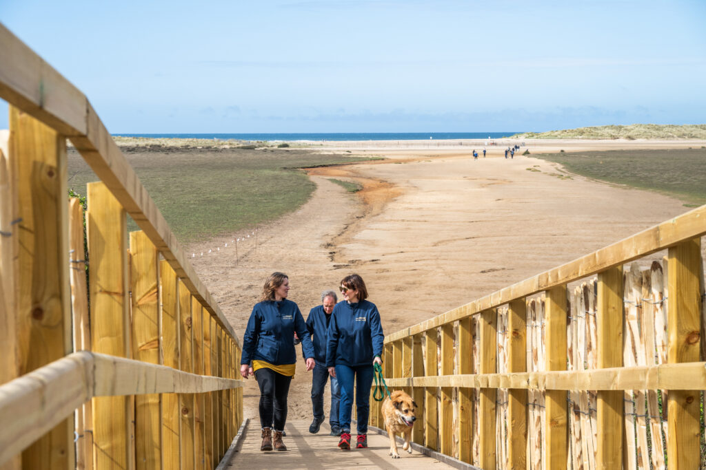 Two women walking up the boardwalk at Holkham. Beside them is wooden handrails. Behind them is a view of Holkham beach.